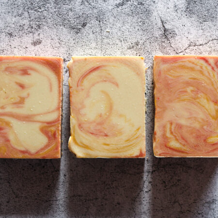 Three bars of soap against a stone like background. The soap is a creamy tan, with swirls of yellow and peach colour throughout.