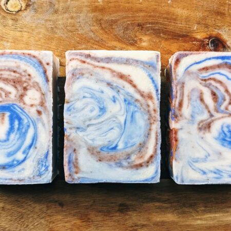 Bars of soap against a wooden board. The soap is white, blue, and speckled red/brown swirls.