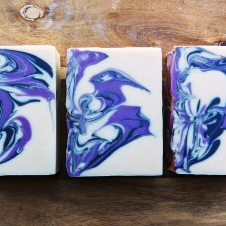 Three bars of soap on a wooden board. The soap is white, with purple and black swirling.