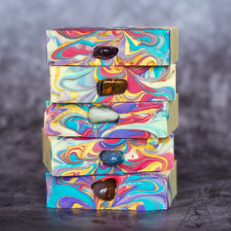 Five bars of soap stacked on top of each other. They're swirled with white, blue, purple, pink, and gold, and contain a tumbled gem pressed into the centre of each bar.