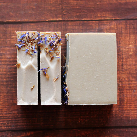 Top down photo of grey creamy bars of soap, on the left showing the top of the soap with cornflower petals and some texturing, and the right soaps, showing the whole bar.