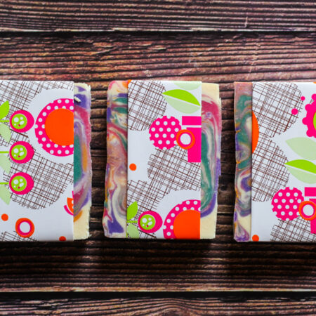 Three bars of brightly coloured and swirled soap, against a wood style background. It's wrapped in bright paper, pink pots and leaves and things.