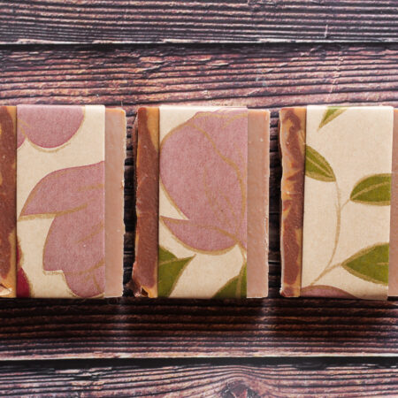 Top down photo of three soaps against a wood looking background. The soap is a creamy brown colour, wrapped in brown paper with a washed out red and green floral pattern.