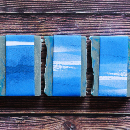 Three bars of blue grey soap against a wood style background. The soap is wrapped in blue and white paper.