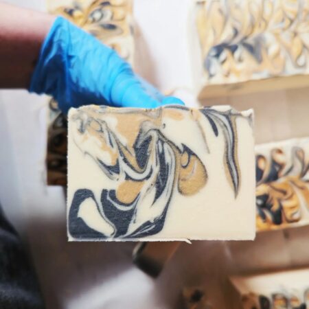 A blue gloved hand holding a creamy white soap, which has swirls of dark grey and gold throughout