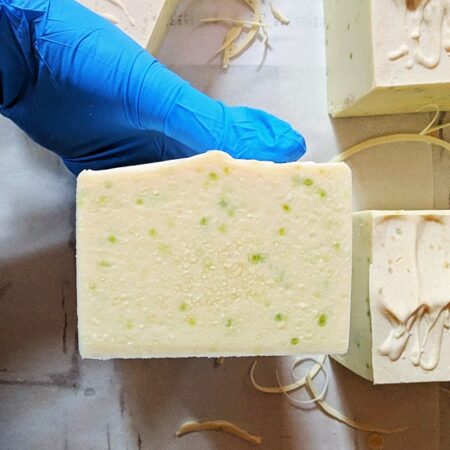 My gloved hand holding freshly cut cucumber soap bars. The soap is a pale creamy yellow with light green speckles.