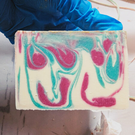 My blue gloved hand holding a freshly cut bar of soap. The soap is white, with swirls of bright green and pink.