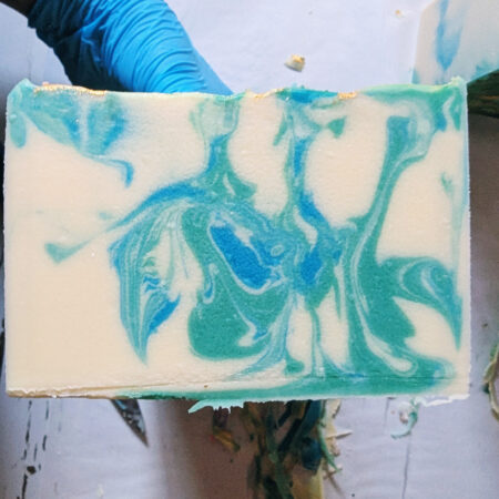 My blue gloved hand holding freshly cut bars of soap. The soap is a creamy white, with swirls of blue and green throughout.