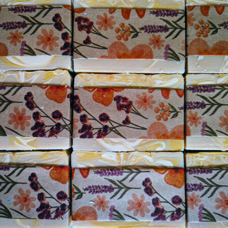 Bars of soap flat laid in their packaging. The soap bars are a creamy brown with swirls of white and yellow. They're in bran paper bands with orange and purple flowers.