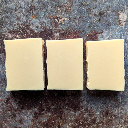 Three bars of creamy white soap flat laid on a metal style background. They are very wonky little rectangles, slightly squashed in. Whoops!