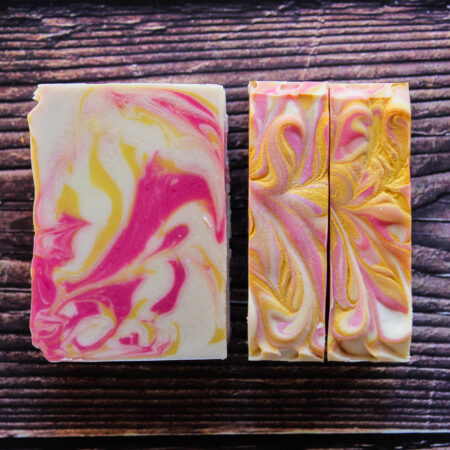 Bars of soap against an apparent wood grain background. The soap is set so you can see two bars tops, and the interior of another bar. The soap is white, and brightly swirled with pink and yellow inside, with gold accents on the top.