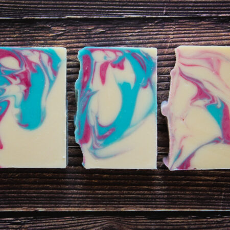 Three bars of soap laid flat against a wood grain background. The soap is white and it's brightly swirled with aqua blue and pink.