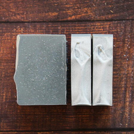 Deep grey bars of soap. They're set against a wood background so you can see the interior of one bar, and the tops of the other two. The tops have greyish white soda ash covering them, and the bar itself has lots of air bubbles visible.